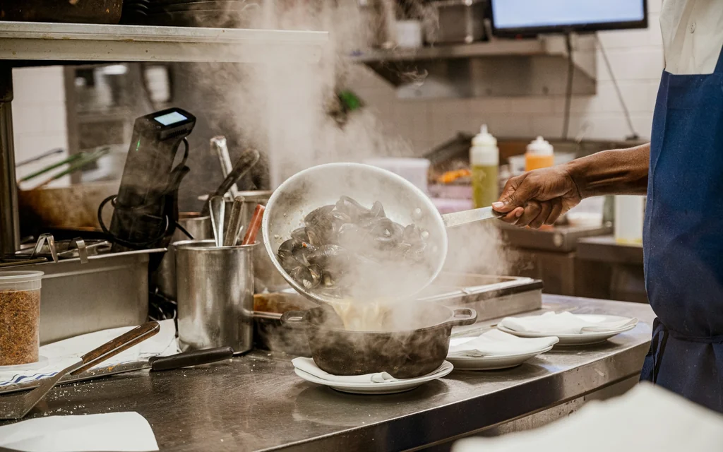 Koch hebt in einer Gastronomieküche eine dampfende Pfanne über die Anrichte