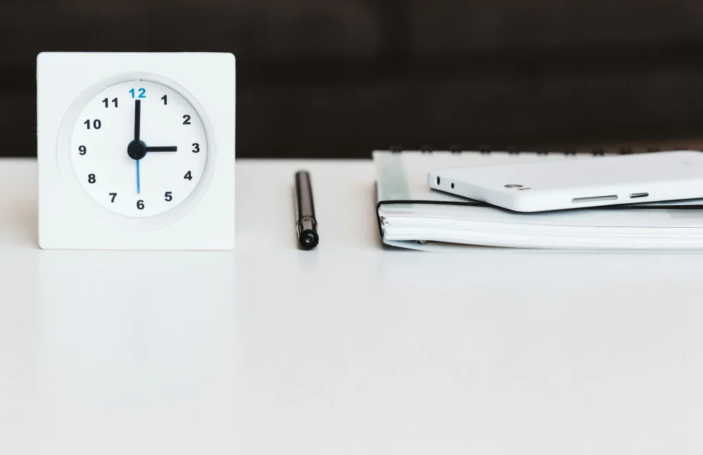 Weisse Tischuhr neben Stift, Notizbuch und Smartphone auf einem hellen Schreibtisch