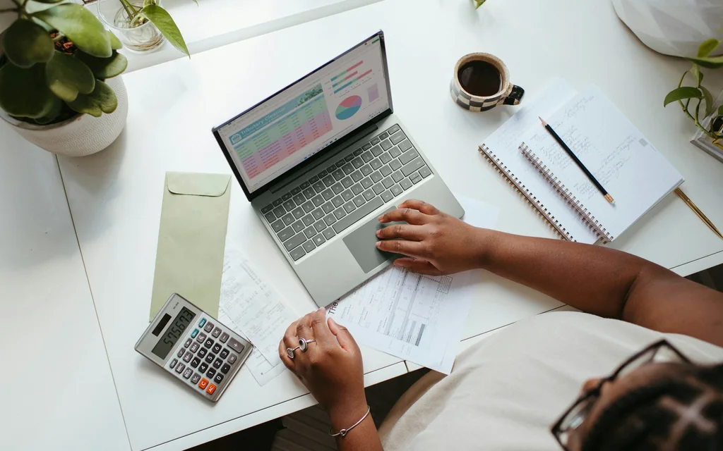 Mitarbeiterin am Laptop mit Taschenrechner, Belegen und Notizen auf dem Schreibtisch